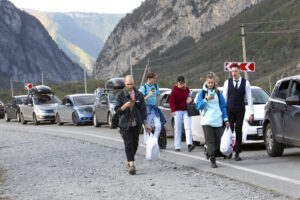 A group of several people walking along the side of a road full of stopped cars. They are carrying bags.