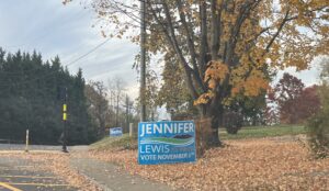 A campaign sign for Jennifer Lewis stands next to a tree and a sidewalk.