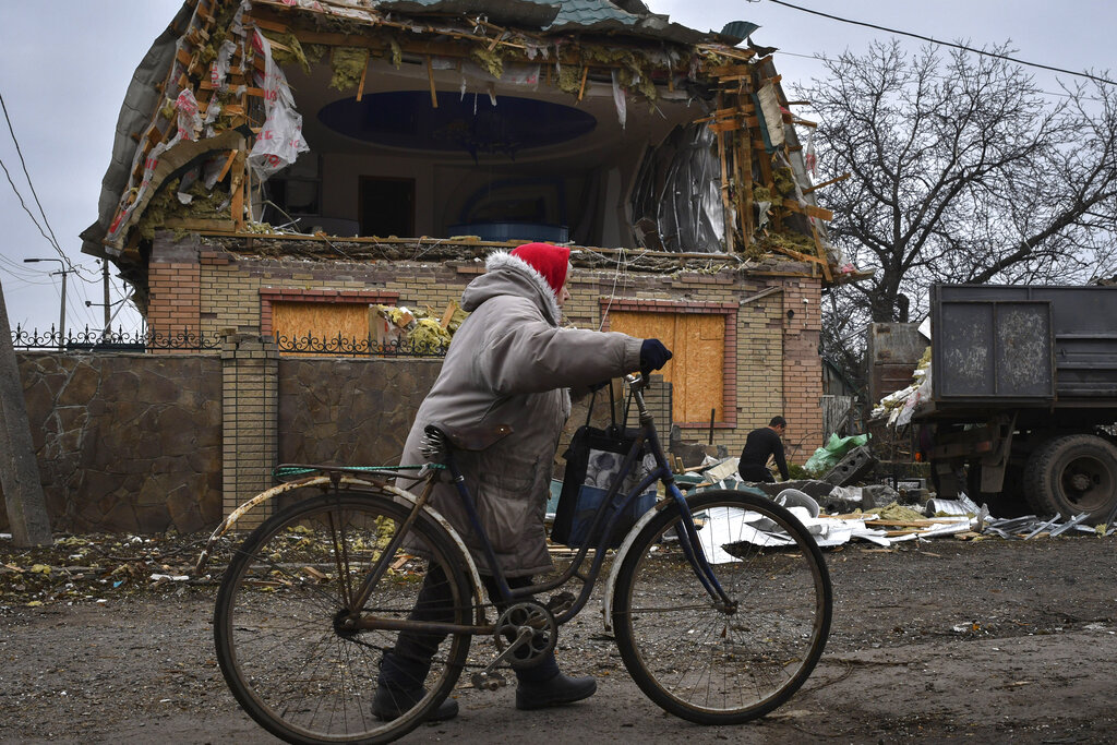 A woman walks a bicycle on a road by a house with a large hole in its second floor.