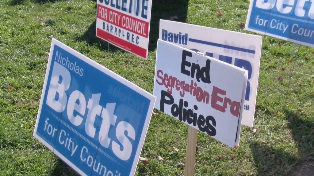 Two signs read "Nicholas Betts for city council" and "end segregation era policies." other signs are around them.