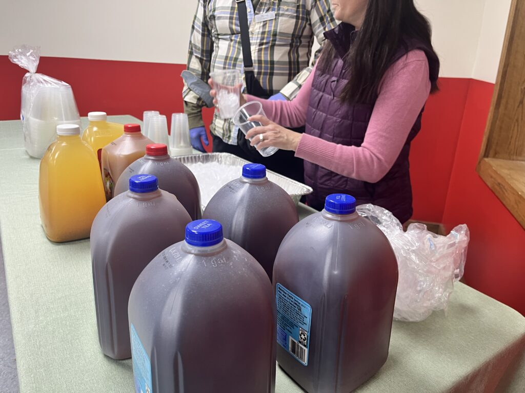 People setting up cups with ice next to gallons of tea and juice jugs