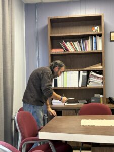 White man with brown beard and shirt, looks over papers above a table and two red chairs
