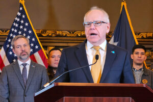 White old man speaking in suit and tie at a podium. American flag, Minnesota flag, and 3 others in the distance standing behind him 