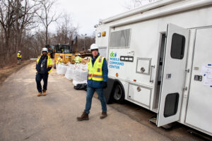 Worker in neon yellow vest and white hat walks in front of large white D.C. Water mobile command center vehicle. Another worker in the distance. 