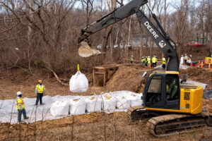 Two workers in neon yellow, machinery lifting large white containers of material, multiple of these on the ground
