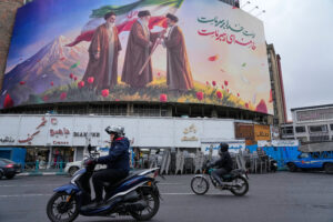 Large jumbo screen depicts late supreme leader passing the Iran flag to his son with a saying in Farsi. Two motobikes pass by as it is in downtown Tehran, Iran.