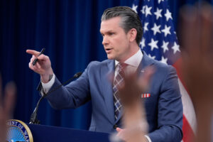 American flag in the background of Pete Hegseth speaker in a suit pointing his finger to somewhere we cant see in the picture.